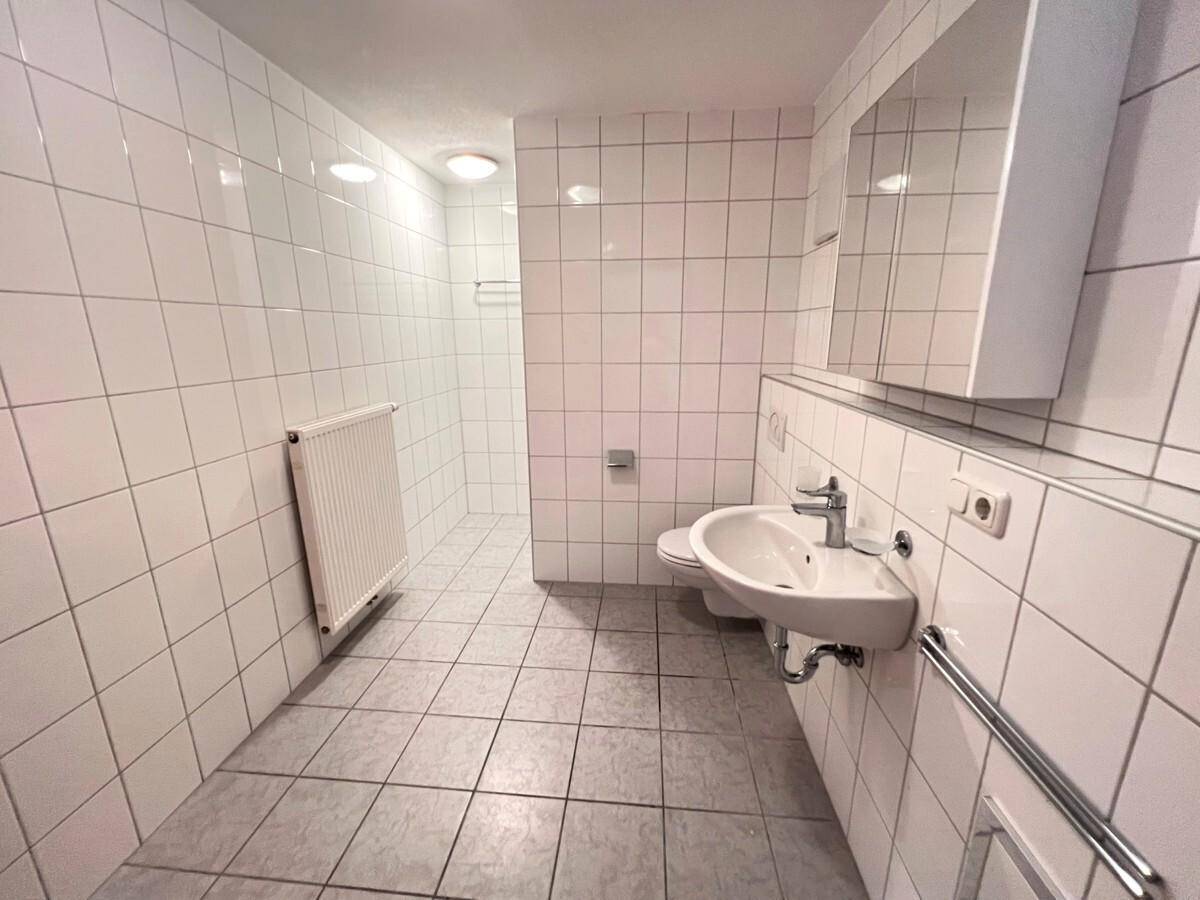 Geräumiges Badezimmer mit weißen Fliesen, Waschbecken und vorbereitetem Duschbereich.