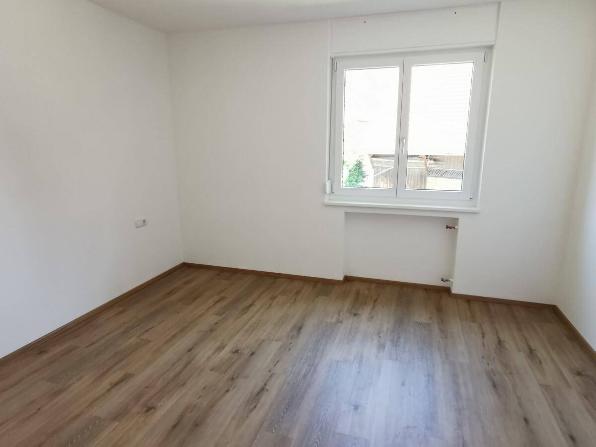Leeres Zimmer mit Laminatboden und einem Fenster, das viel Tageslicht hereinlässt.
