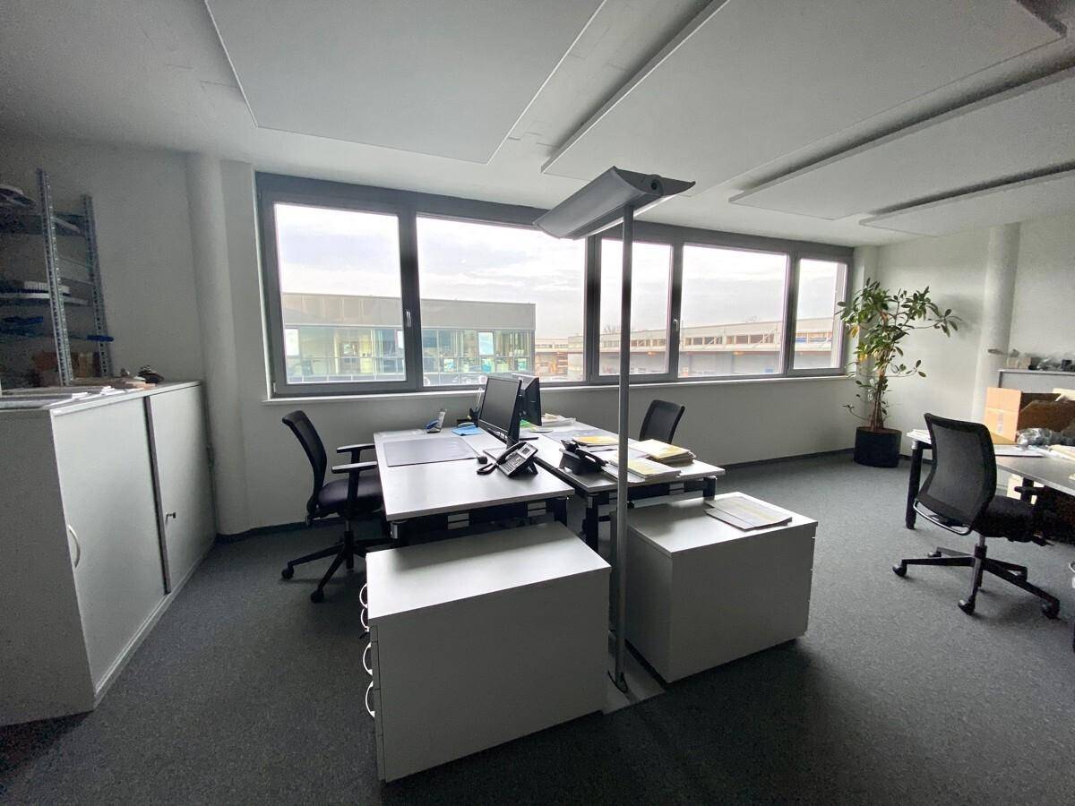 Helles Büro mit grauem Teppichboden, großen Fenstern und moderner Büroeinrichtung.