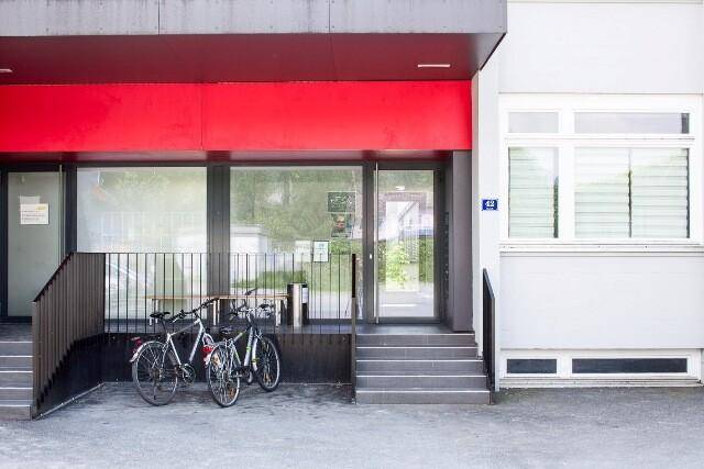 Eingangsbereich eines modernen Gebäudes mit Treppen, Geländer und abgestellten Fahrrädern vor der Glasfassade.