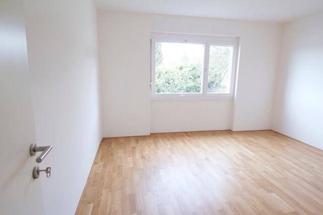 Leeres Zimmer mit hellem Parkettboden und einem Fenster mit Blick ins Grüne.