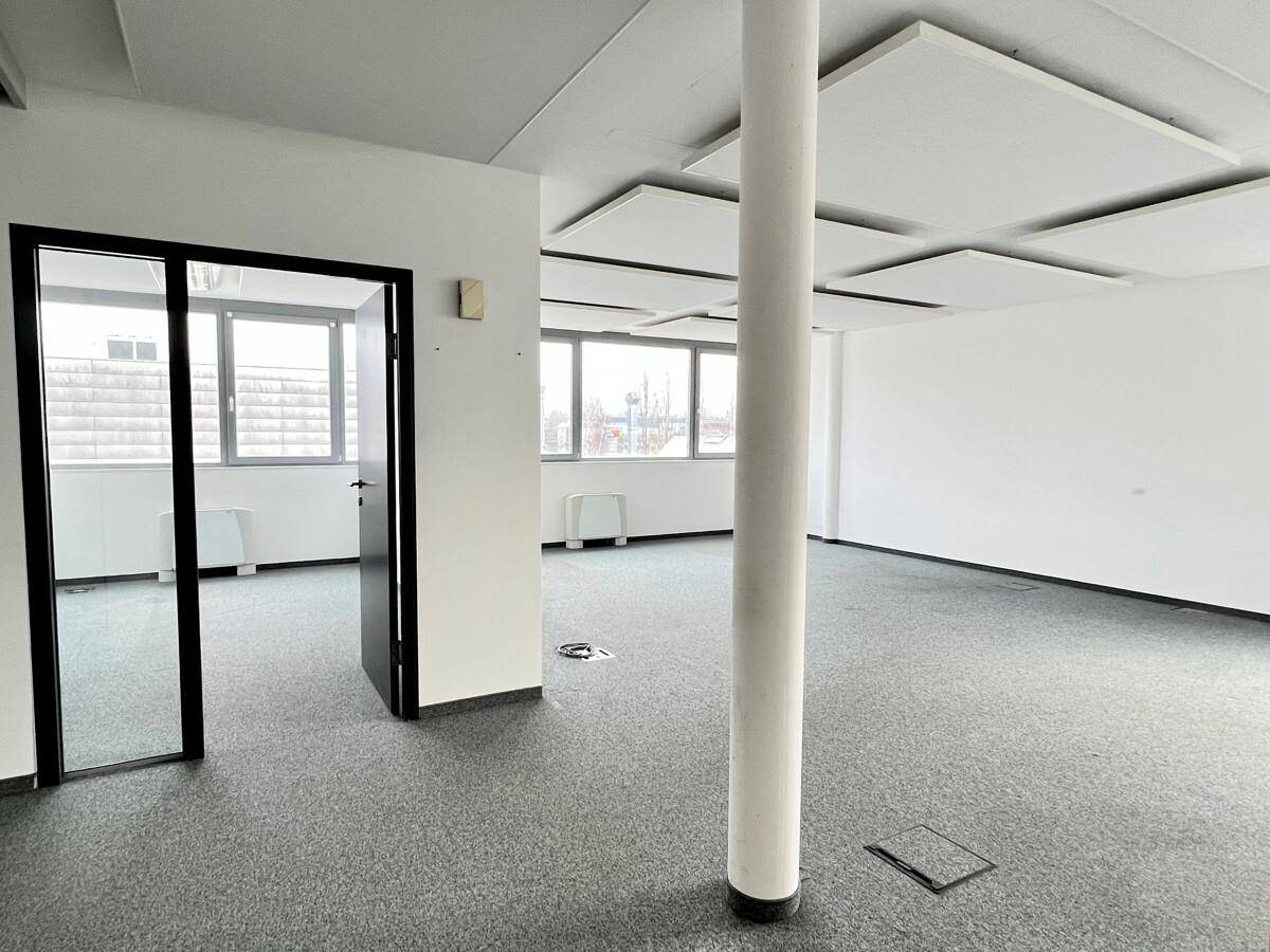 Großes, offenes Büro mit zentraler Säule, grauem Teppichboden, großen Fenstern und schallabsorbierenden Deckenplatten.