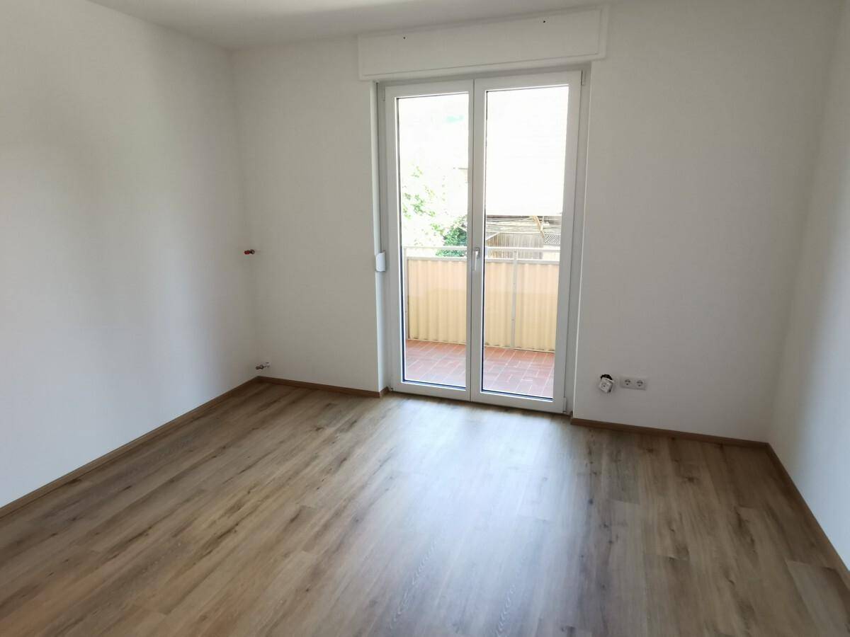 Helles, leeres Zimmer mit Laminatboden und Zugang zu einem Balkon.