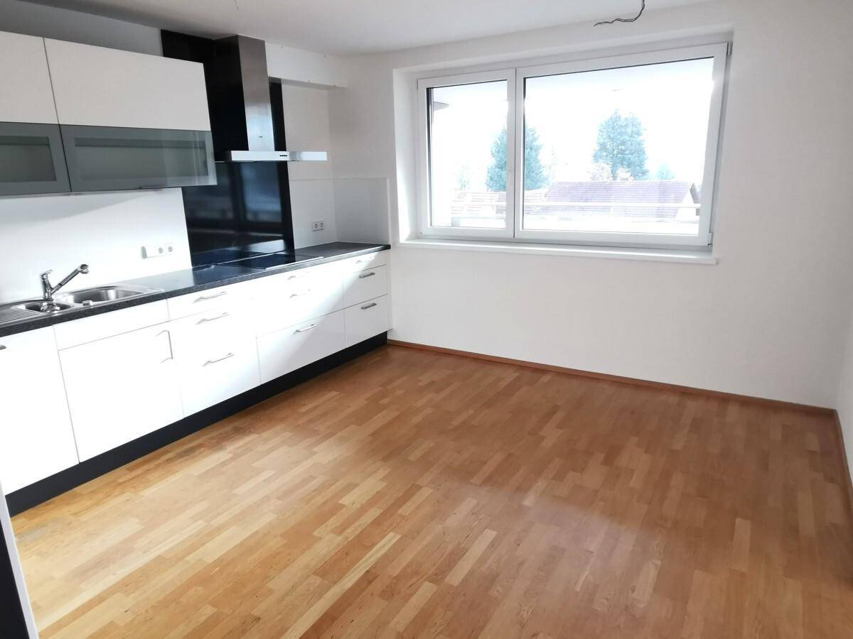 Moderne Küche mit weißen Schränken, dunkler Arbeitsplatte und Parkettboden, hell durch ein großes Fenster.