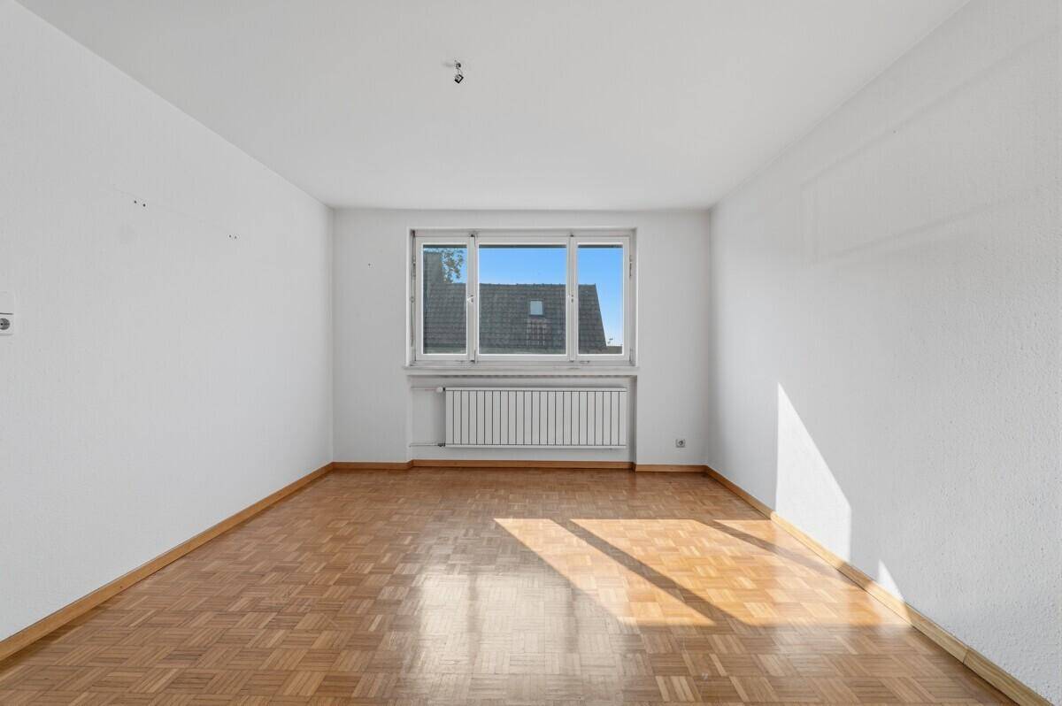 Heller, leerer Raum mit Parkettboden und großem Fenster, das viel Tageslicht hereinlässt.