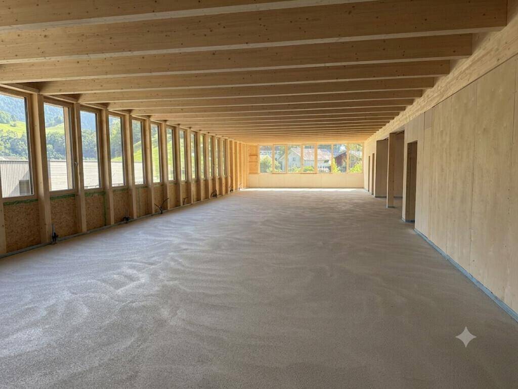 Geräumiger, heller Innenraum mit sichtbaren Holzdeckenbalken und großen Fensterfronten mit Bergblick.