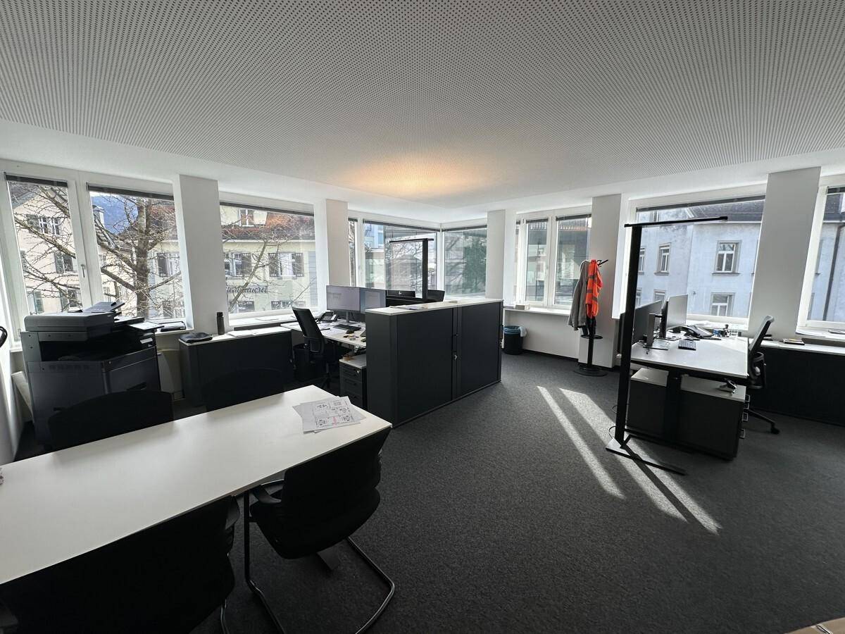 Geräumiges Büro mit mehreren Arbeitsplätzen, großen Fenstern und Teppichboden.