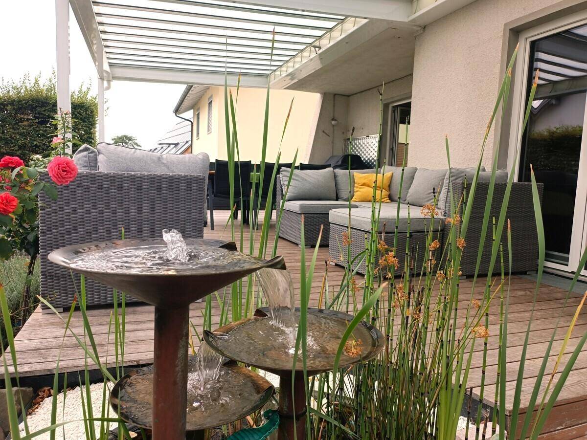 Gemütliche Lounge-Ecke auf der Holzterrasse mit dekorativem Brunnen und üppiger Bepflanzung.