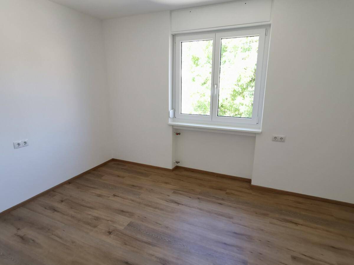 Helles, leeres Zimmer mit Laminatboden und einem Fenster mit Blick ins Grüne.