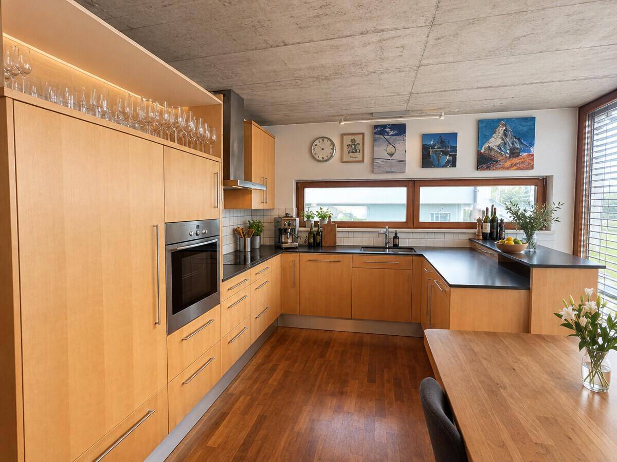 Moderne Küche mit hellen Holzschränken, dunklen Arbeitsplatten und einem Essbereich unter einer Betondecke.