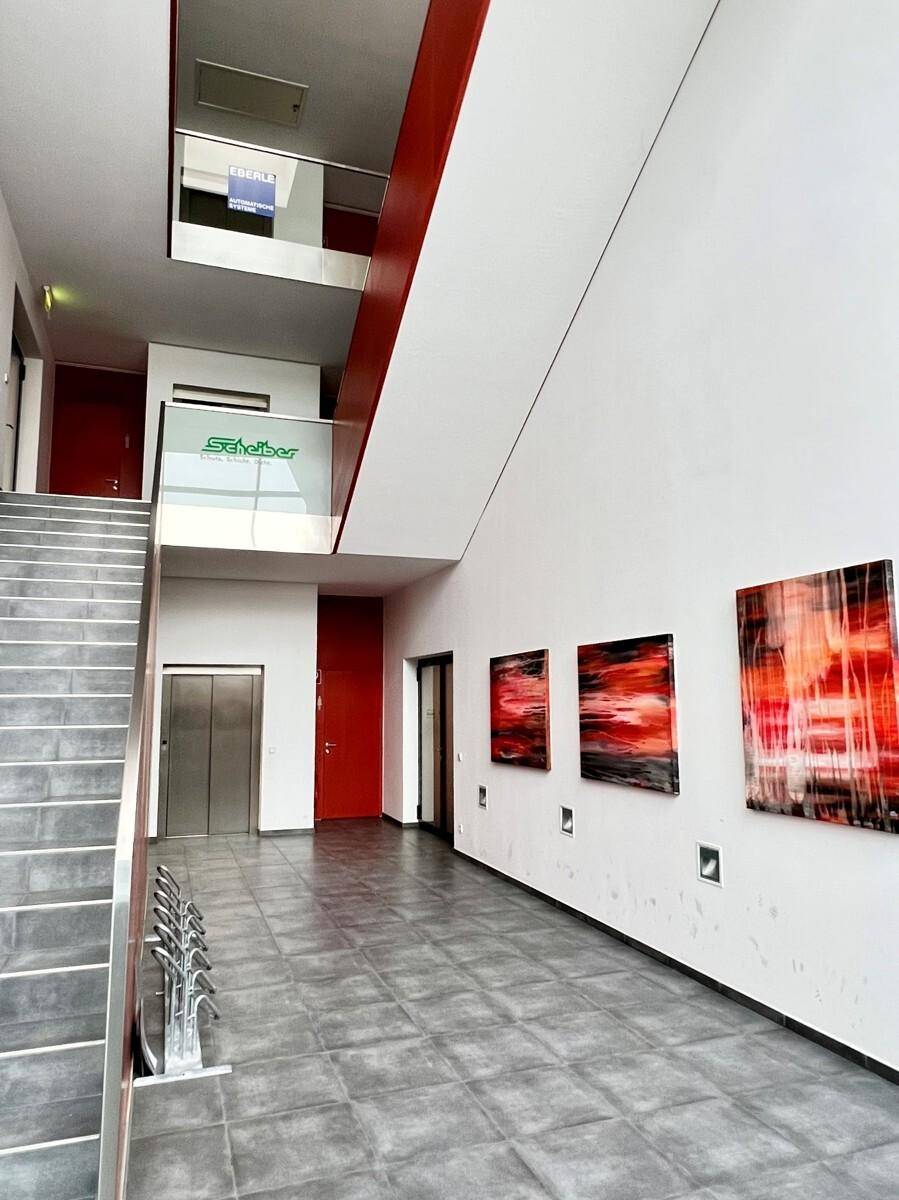 Modernes Foyer mit grauen Fliesen, einer Treppe, einem Aufzug und roten Akzenten an den Wänden.