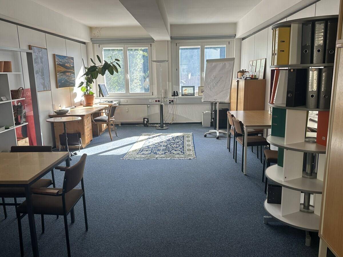 Helles Büro mit blauem Teppichboden, großen Fenstern und Blick ins Grüne, ausgestattet mit Schreibtischen und Regalen.