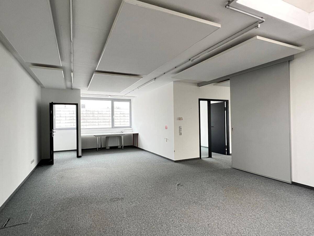 Geräumiges Büro mit grauem Teppichboden, großen Fenstern, schallabsorbierenden Deckenplatten und Zugang zu weiteren Räumen.