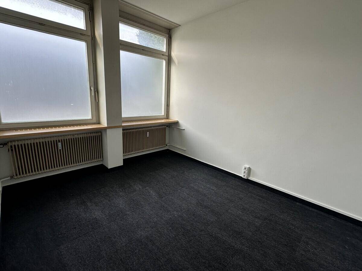 Heller Büroraum mit großen Fenstern, schwarzem Teppichboden und Heizkörpern unter den Fensterbänken.