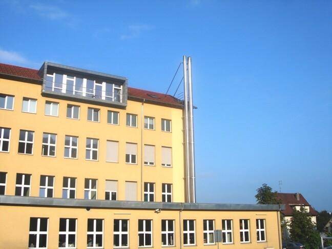 Gebäudefassade mit zahlreichen Fenstern, einem modernen Dachaufbau und einem hohen Schornstein unter blauem Himmel.