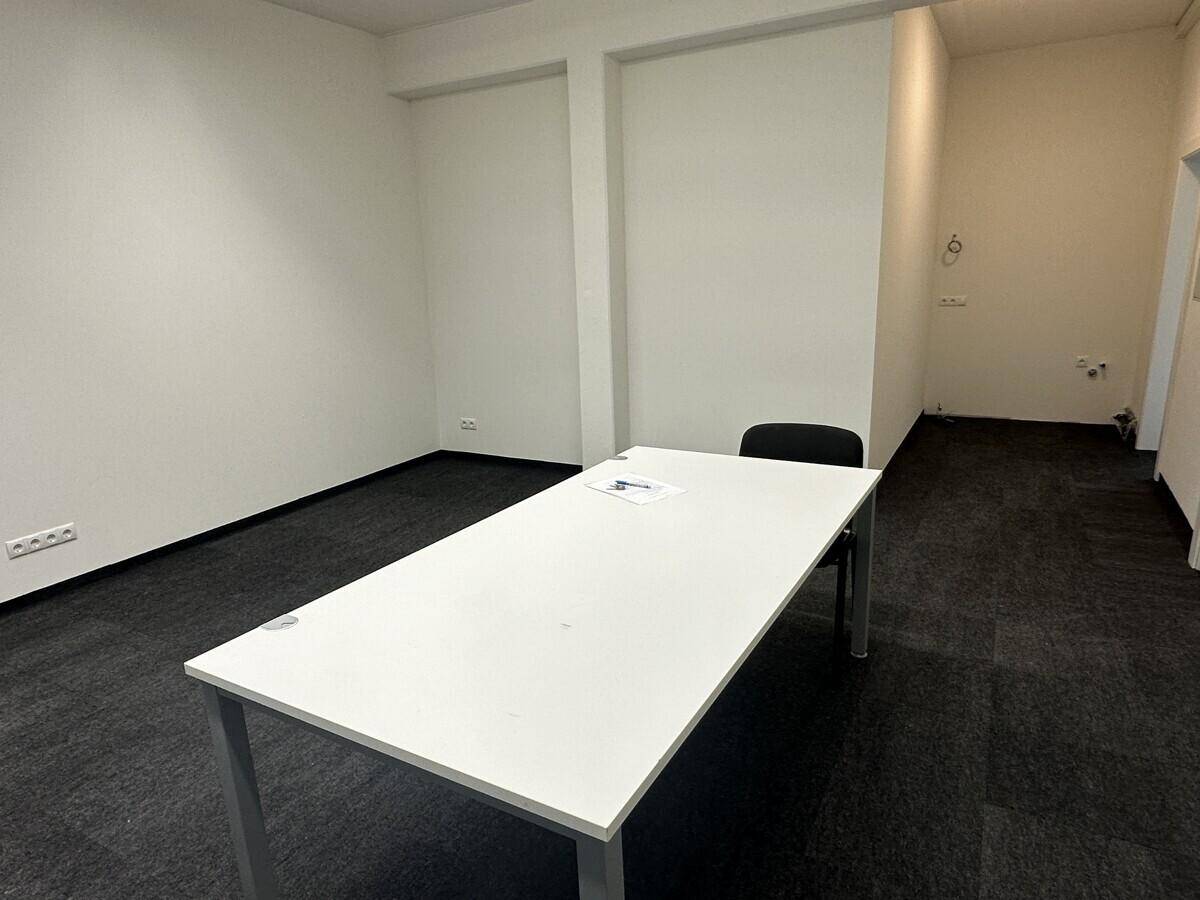 Geräumiger Büroraum mit weißem Tisch, schwarzem Teppichboden und hellen Wänden, bereit zur Nutzung.