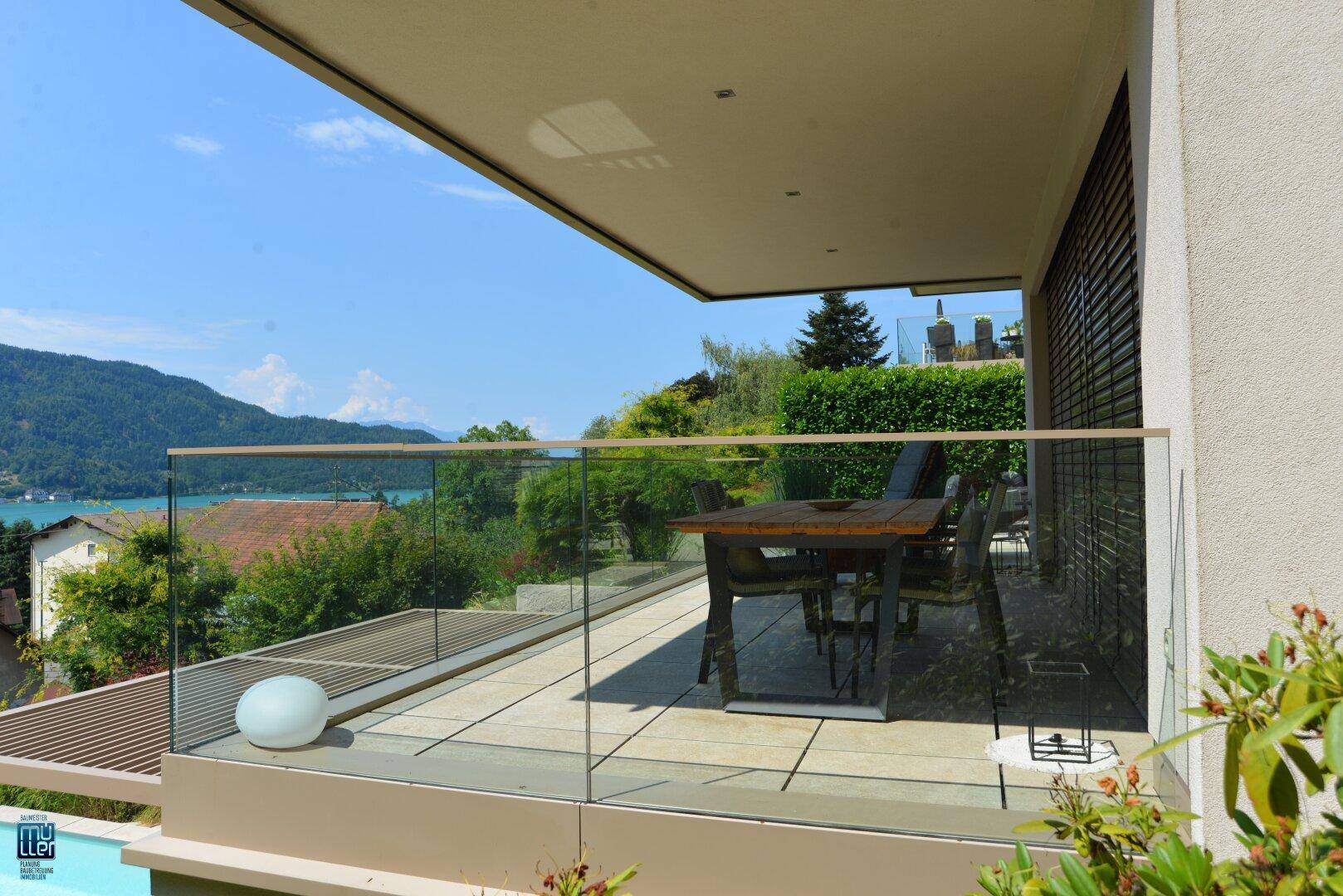 Geräumiger Balkon mit Esstisch und Glasgeländer, der einen weiten Blick auf den See und die Landschaft bietet.