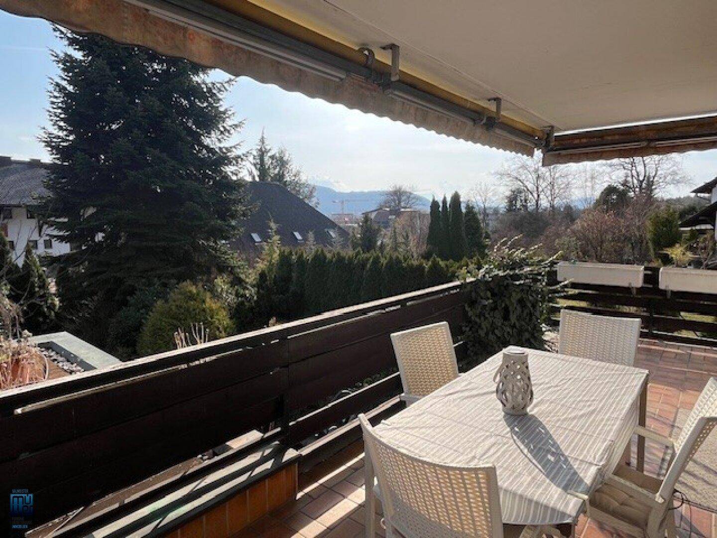 Sonniger Balkon mit Esstisch, Markise und einem Panoramablick auf die grüne Umgebung und Berge.