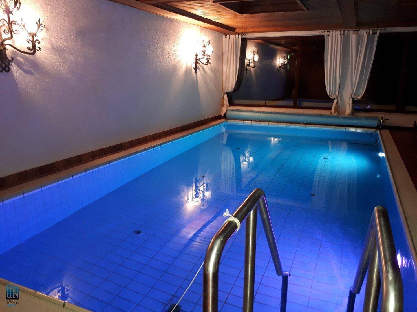 Beleuchteter Innenpoolbereich mit blauen Fliesen und einem direkten Zugang zu weiteren Räumen.