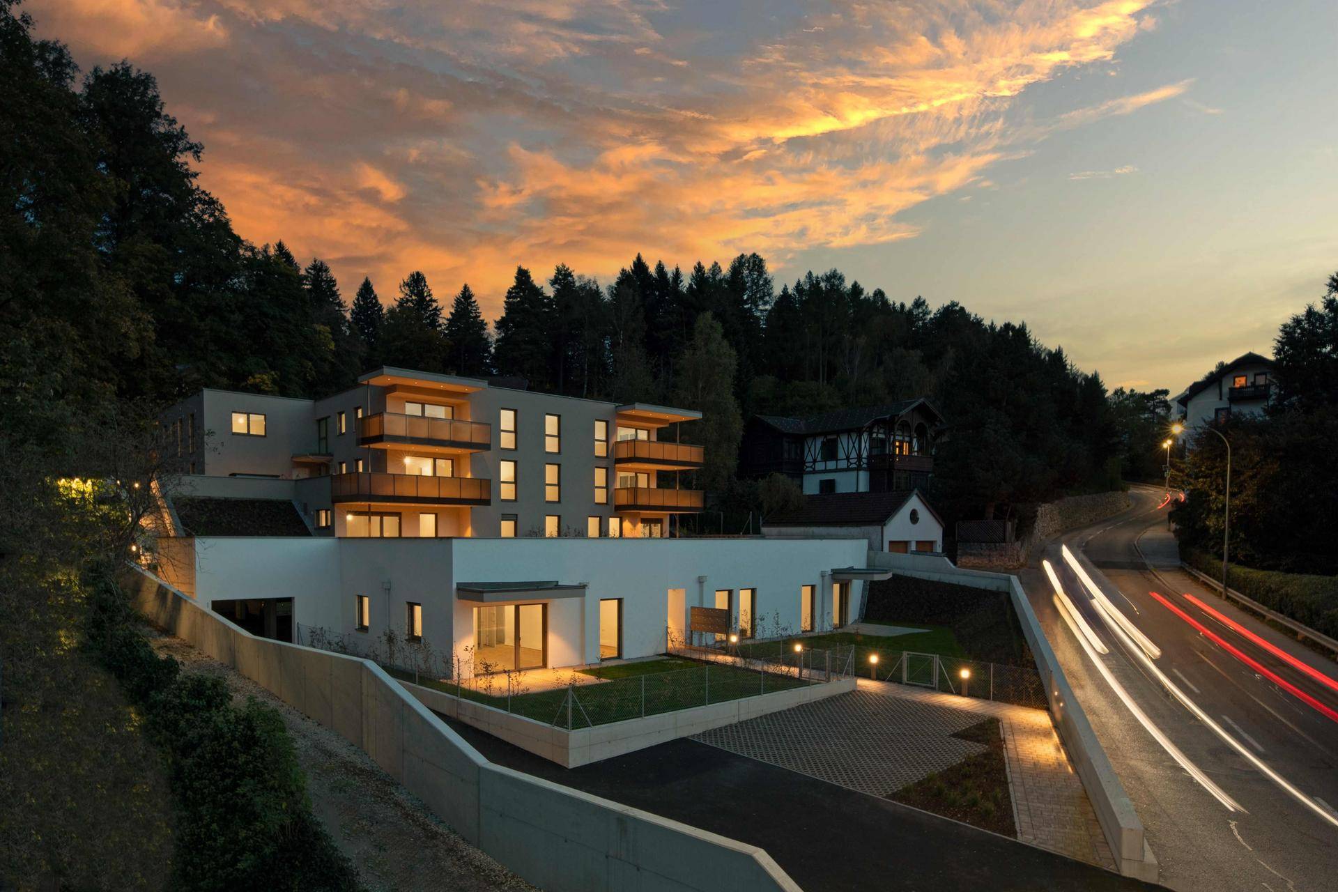 Moderne Wohnanlage bei Sonnenuntergang mit beleuchteter Zufahrt und Balkonen.