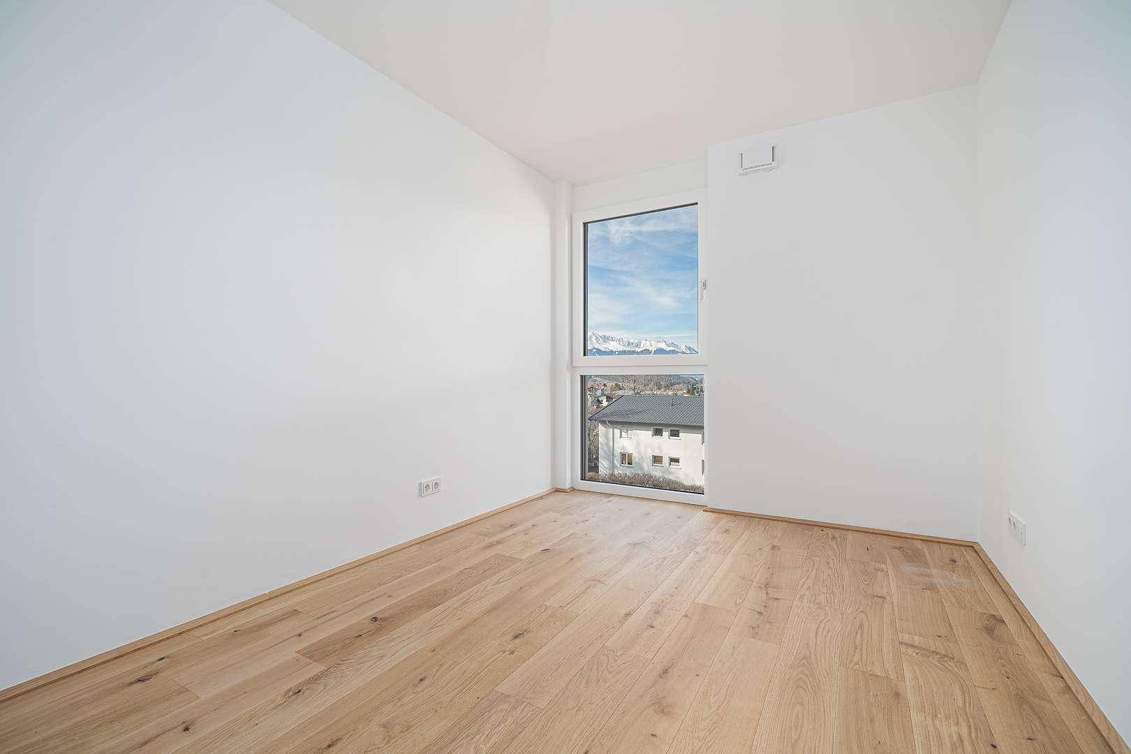 Leeres Zimmer mit Holzparkett und Fenster mit Blick auf die Berge und umliegende Häuser.
