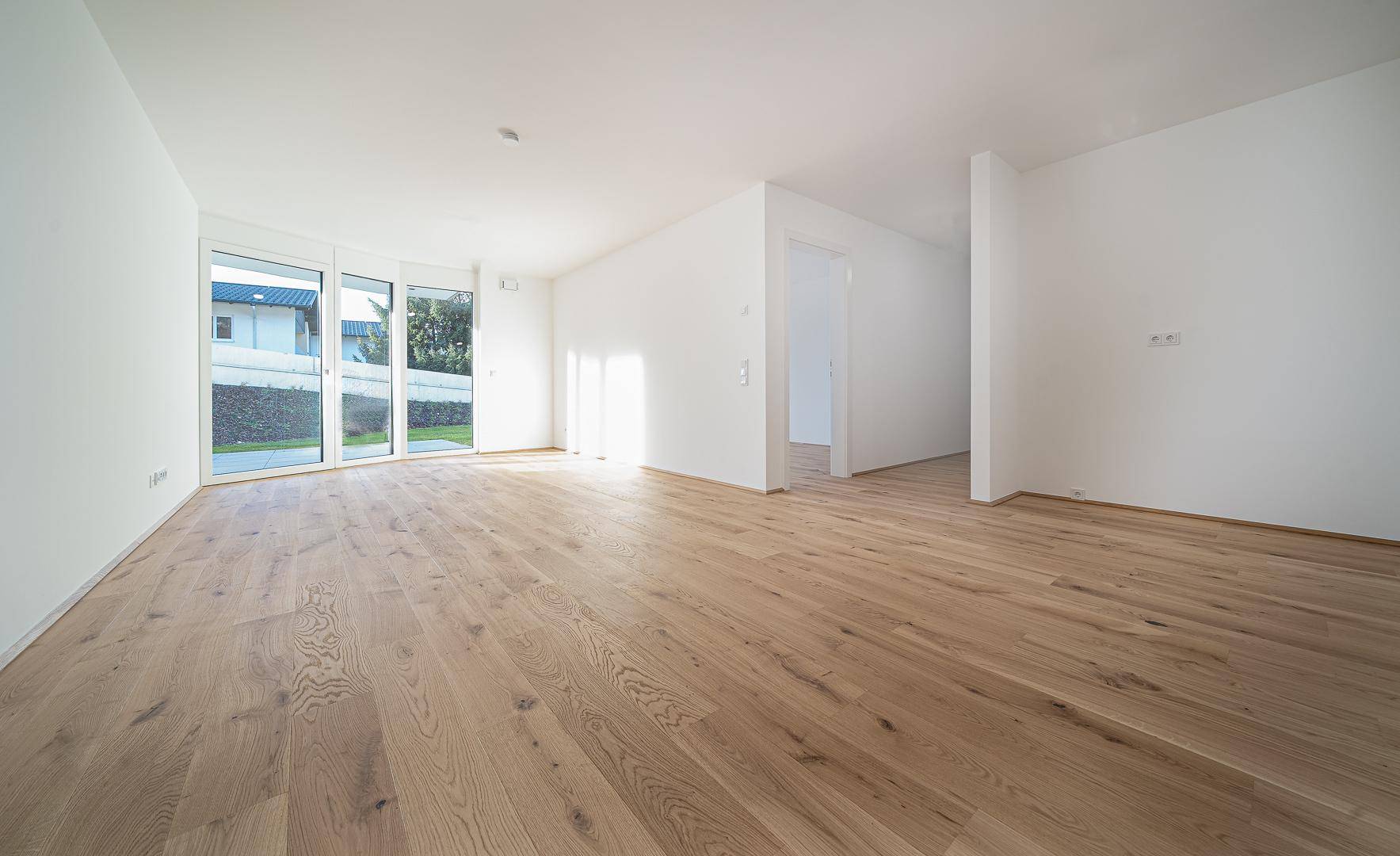 Helles, leeres Wohnzimmer mit Eichenparkettboden und großen Fenstern zum Garten.