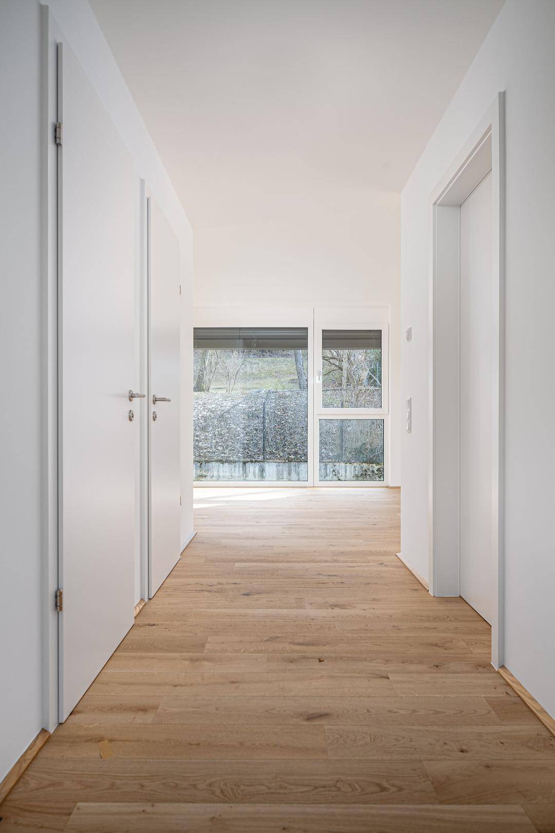 Heller Flur mit Holzboden und weißen Türen, führt zu einem lichtdurchfluteten Raum.