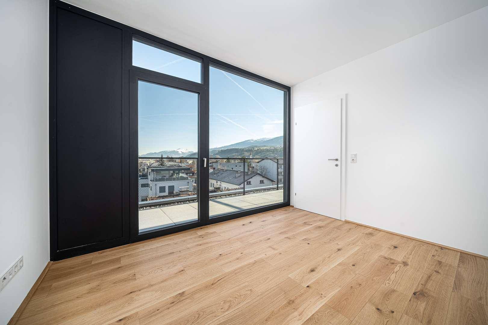 Helles Zimmer mit Holzboden, großen Fenstern und Blick auf die Stadt und die Berge.