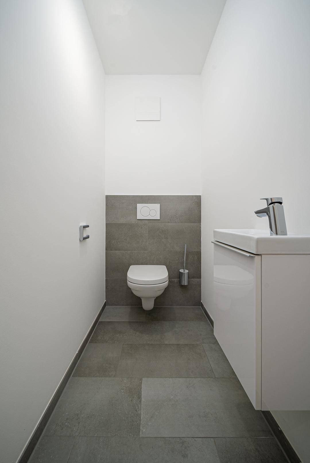 Separates Gäste-WC mit Wand-WC, kleinem Waschtisch und modernen grauen Fliesen.