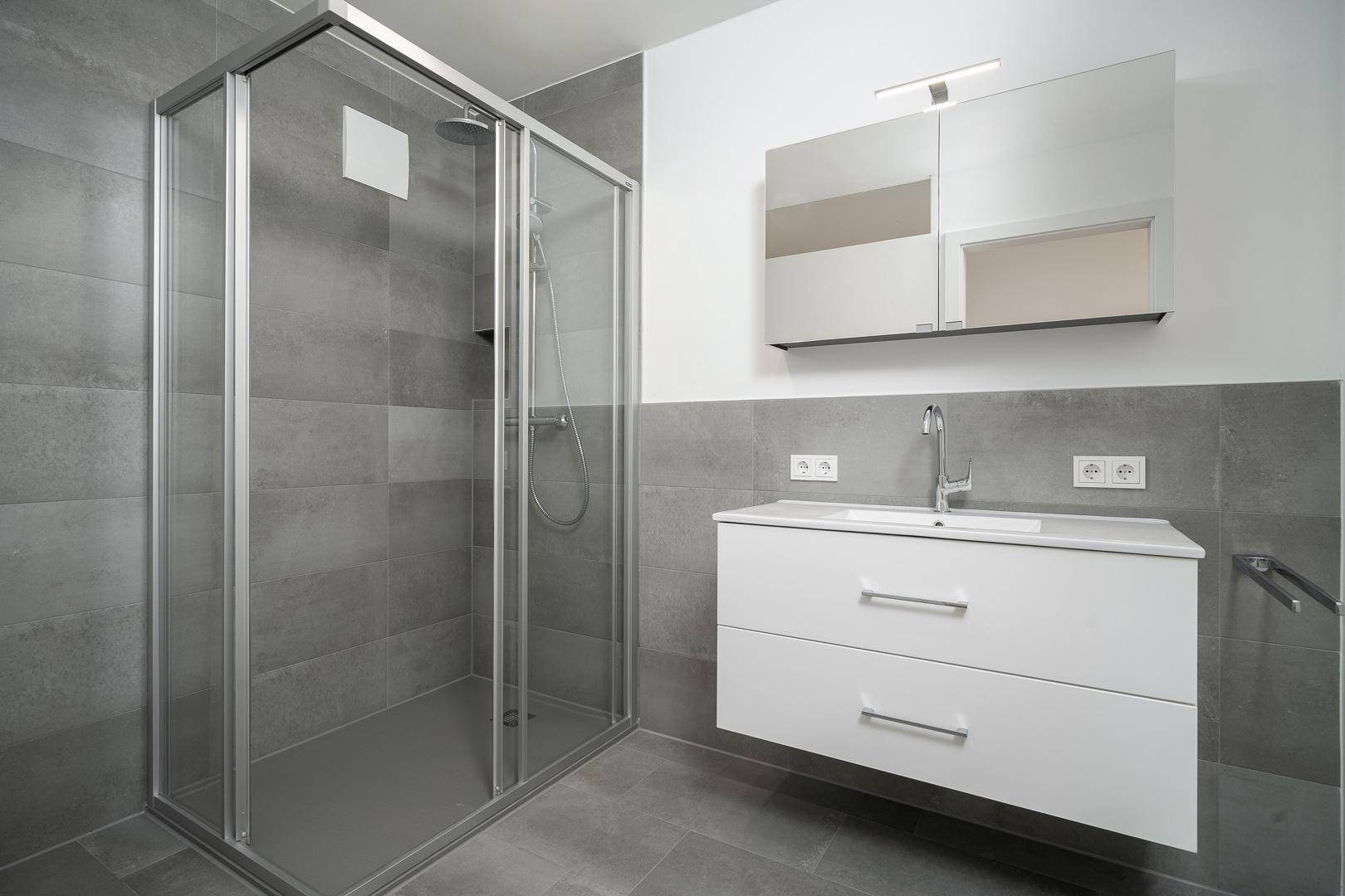 Modernes Badezimmer mit großer Duschkabine, Waschtisch und grauen Fliesen.