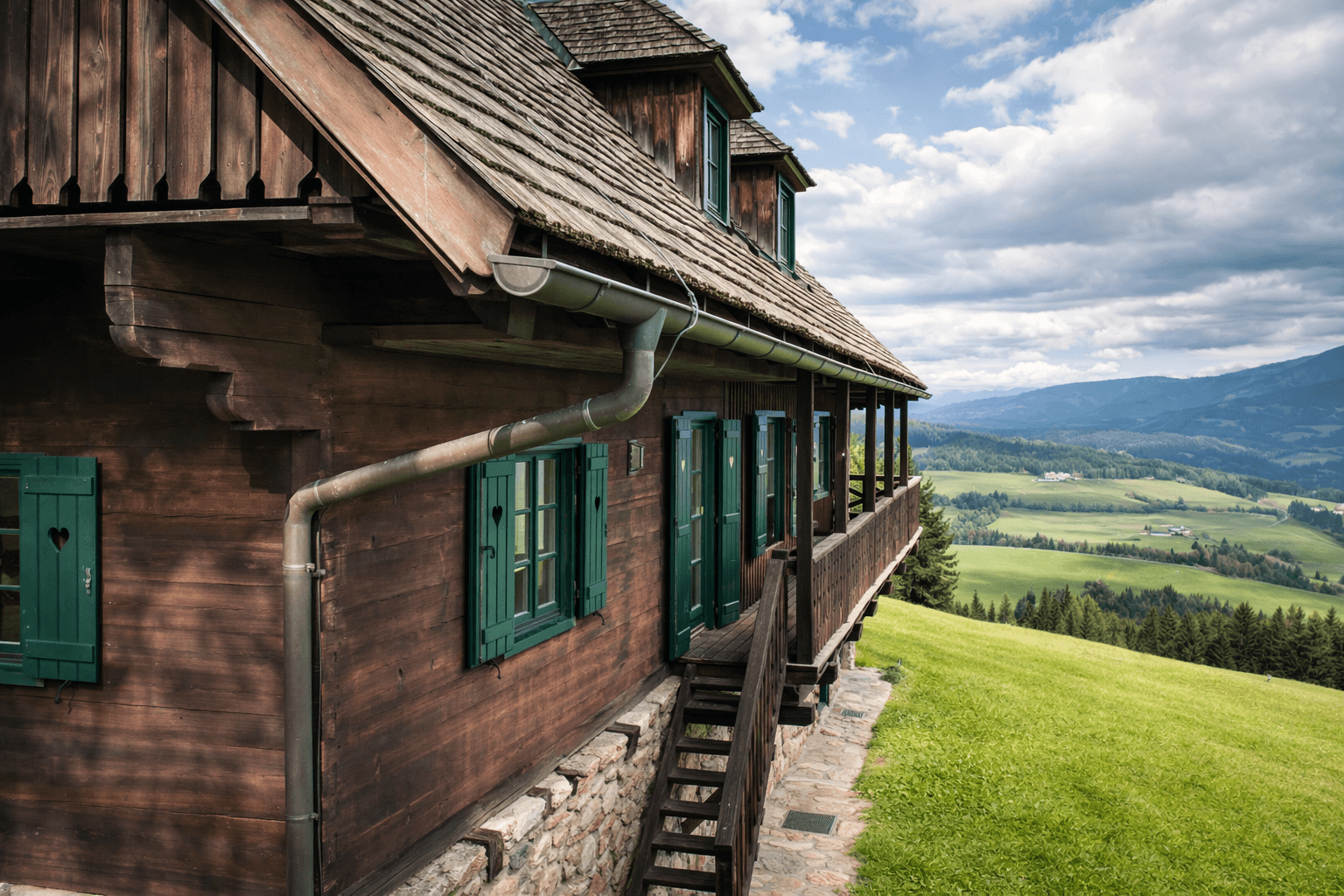 Seitenansicht eines Holzhauses mit Balkon und weitem Blick über die grüne Hügellandschaft.