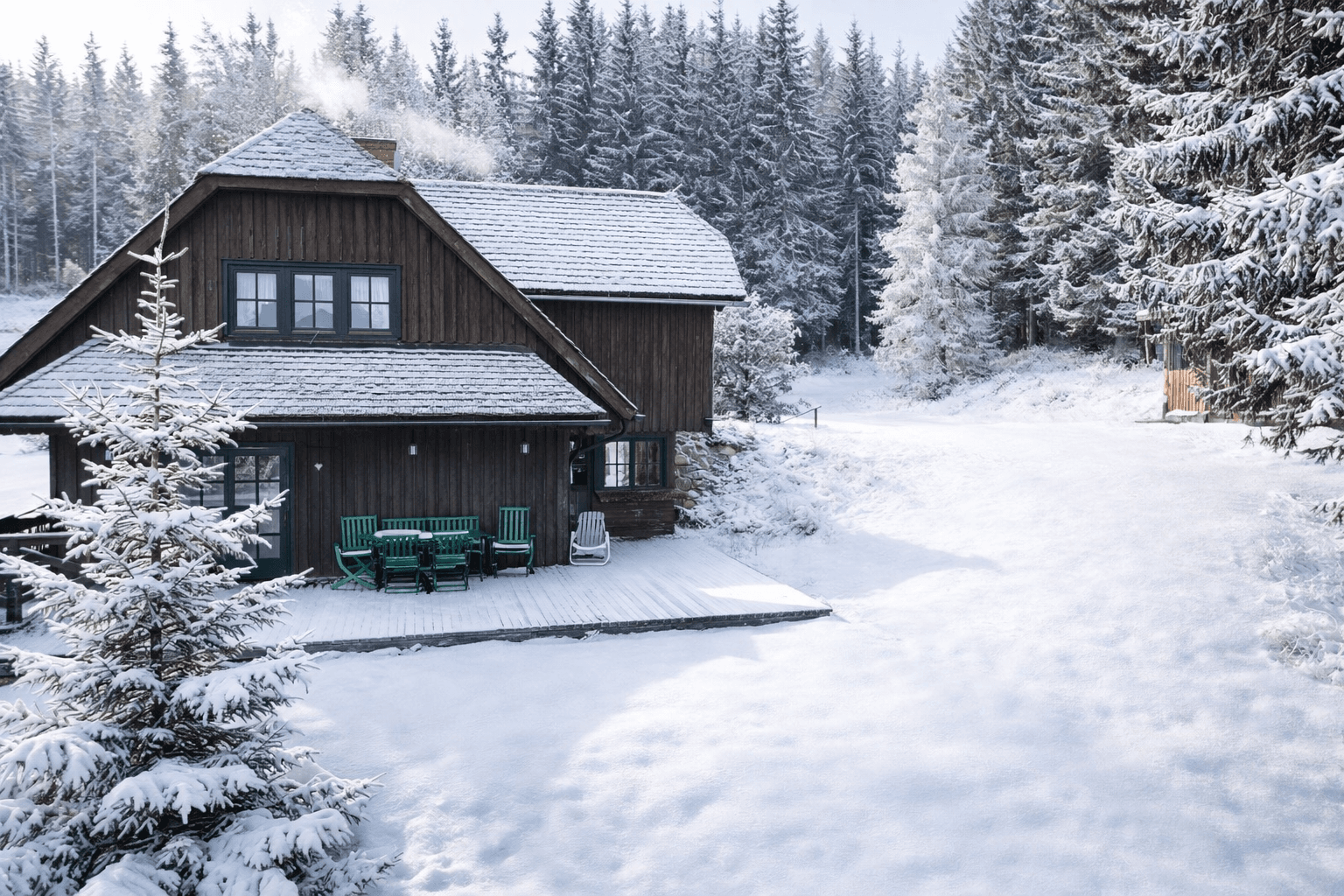Holzhaus mit großer Holzterrasse und Gartenmöbeln, umgeben von verschneiten Bäumen.