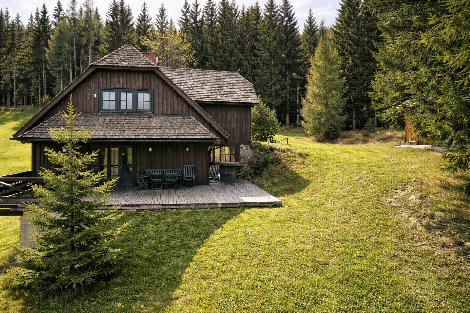 Holzhaus mit großer Holzterrasse und Gartenmöbeln, umgeben von hohen Nadelbäumen.