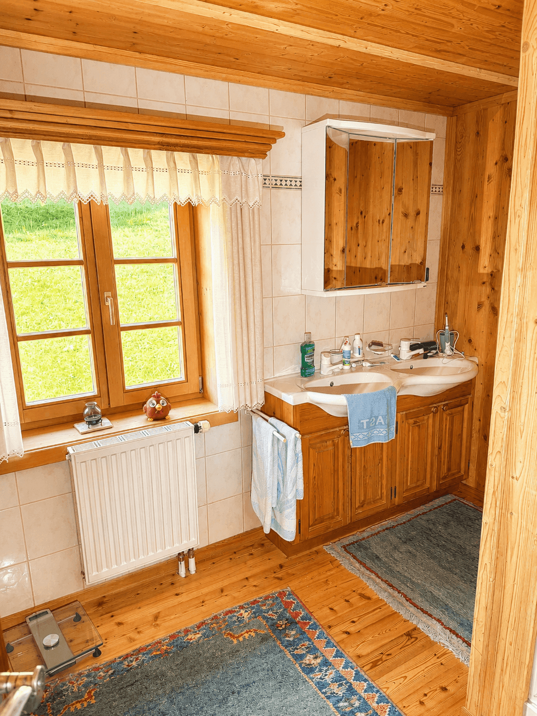 Helles Badezimmer mit Doppelwaschbecken, Holzfußboden und Fenster mit Blick ins Grüne.