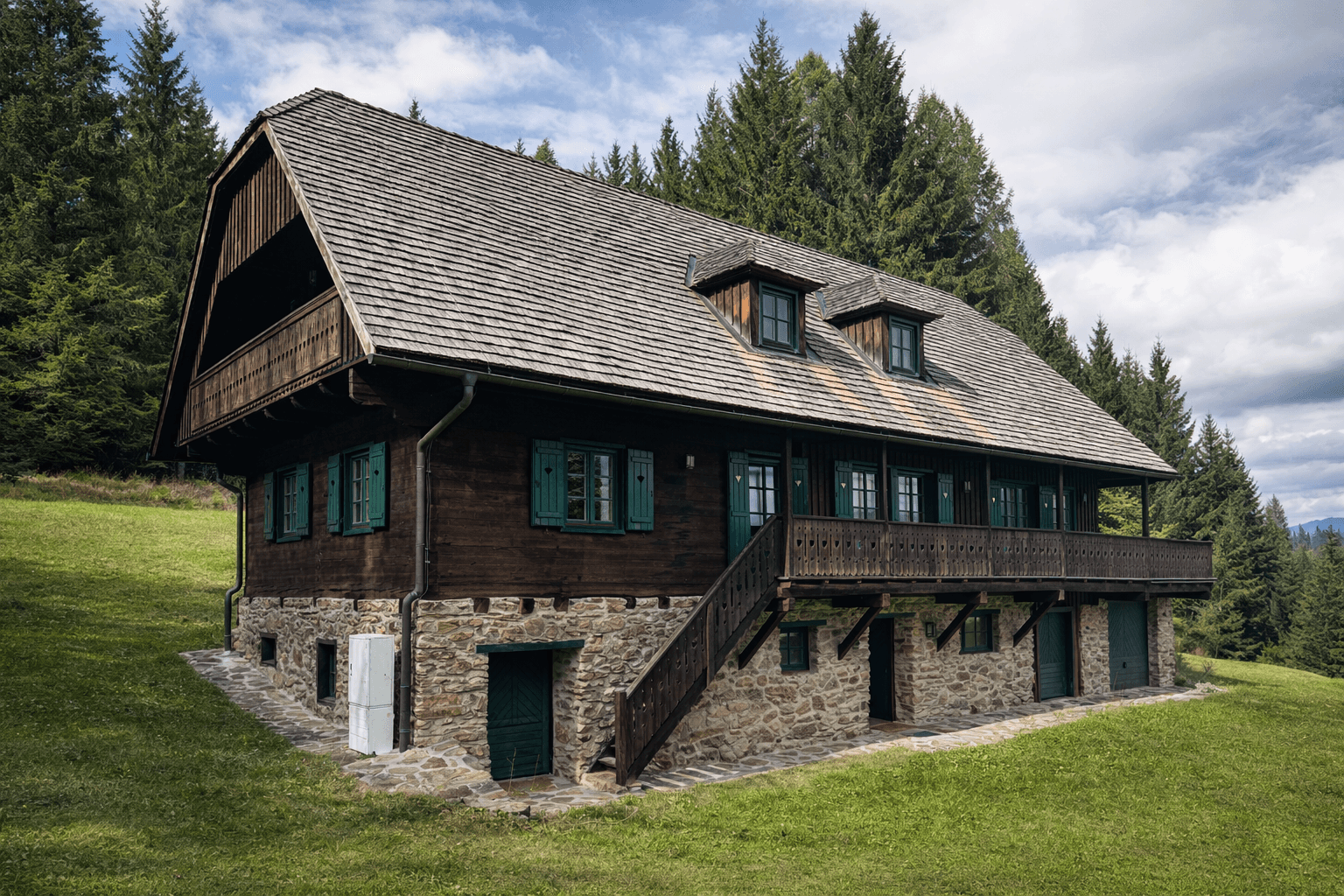 Rustikales Holzhaus mit Steinsockel und Balkon in einer weitläufigen grünen Landschaft.