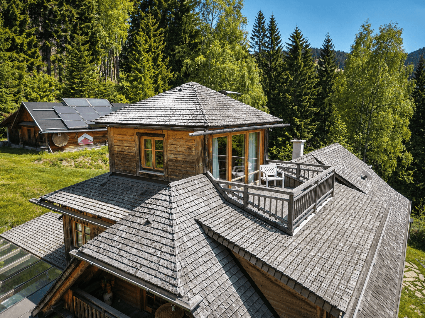 Rustikales Holzhaus mit komplexer Dachkonstruktion und kleiner Dachterrasse inmitten grüner Natur.