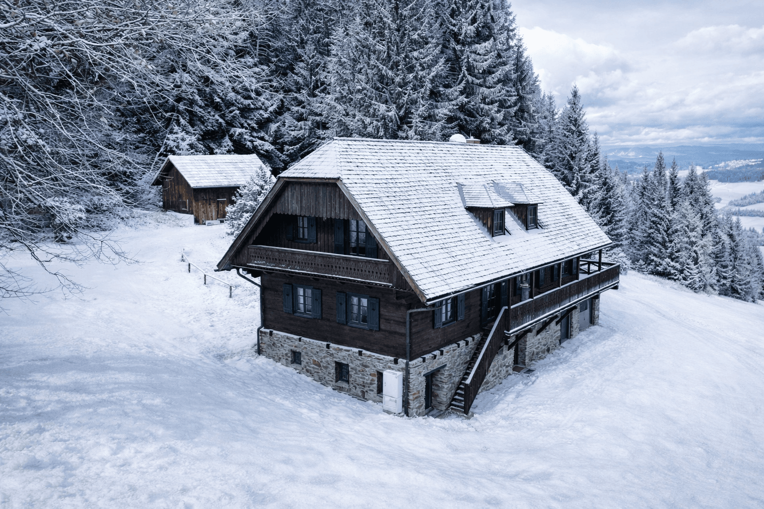 Winteransicht eines traditionellen Holzhauses mit schneebedecktem Dach und umgebendem Wald.