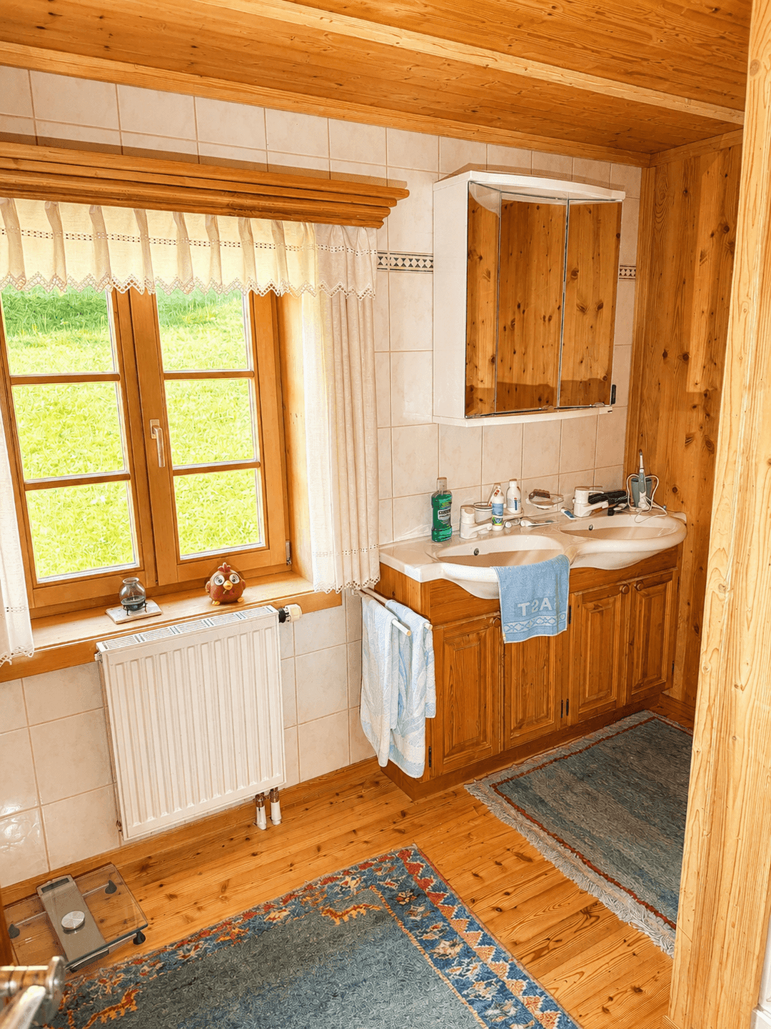 Helles Badezimmer mit Holzvertäfelung, Doppelwaschbecken und Fenster mit Blick ins Grüne.