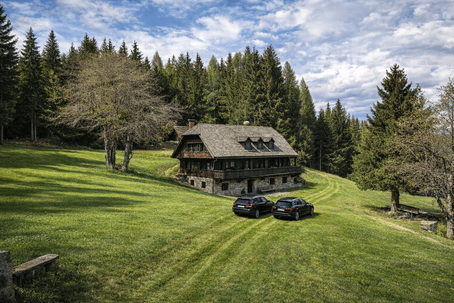 Traditionelles Holzhaus mit Steinfundament in grüner Hügellandschaft mit zwei geparkten Autos.