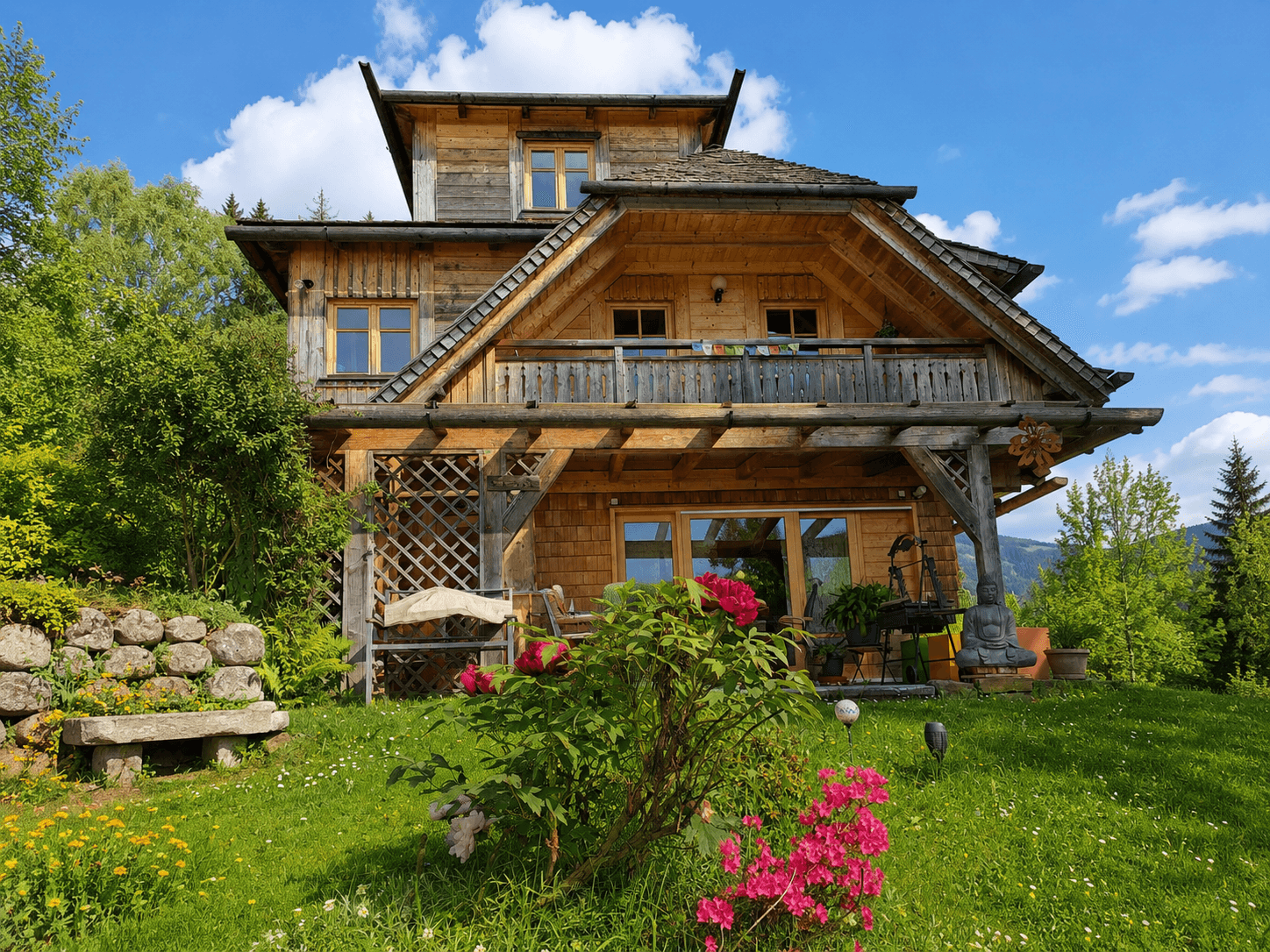 Charmantes Holzhaus mit Balkon und blühendem Vorgarten unter blauem Himmel.