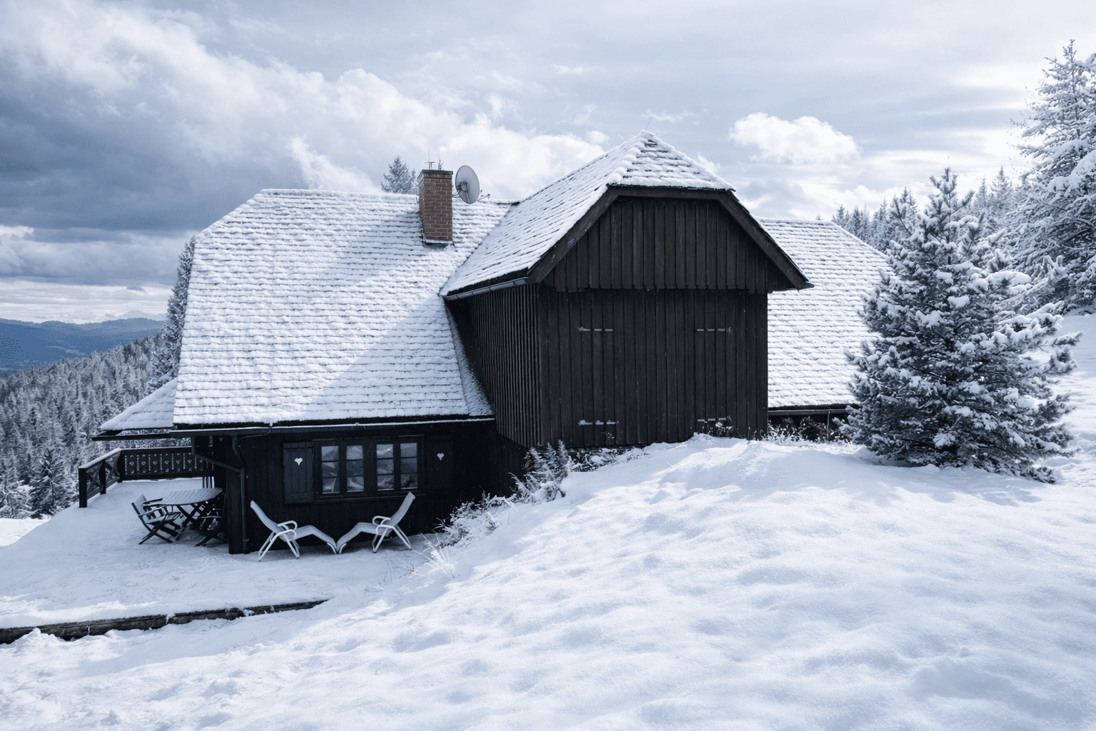 Traditionelles Holzhaus mit schneebedecktem Dach und Terrasse in winterlicher Bergkulisse.