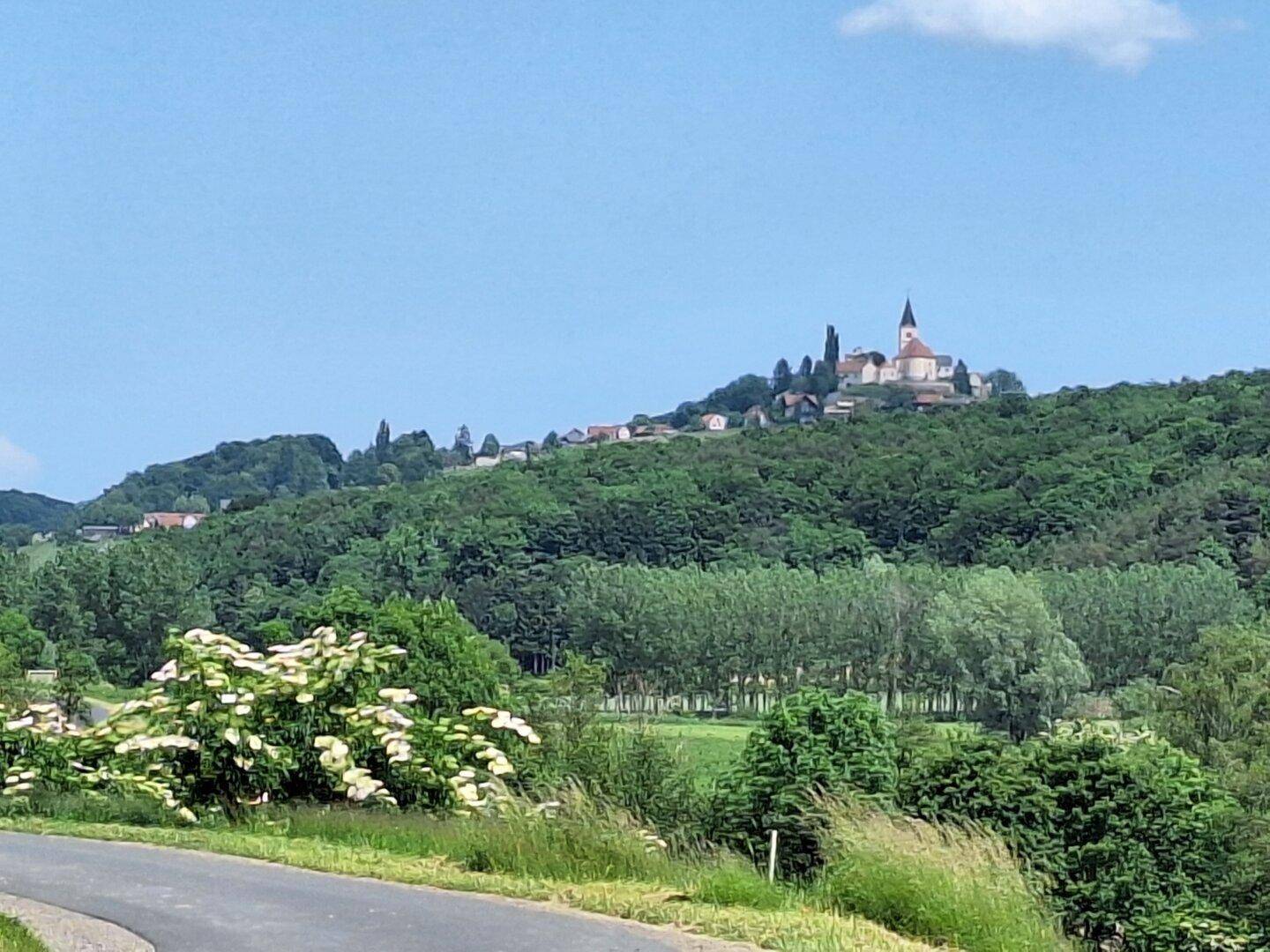 Landstraße im Vordergrund, die zu einem dichten Wald und einem entfernten Hügel mit einer Kirche führt.