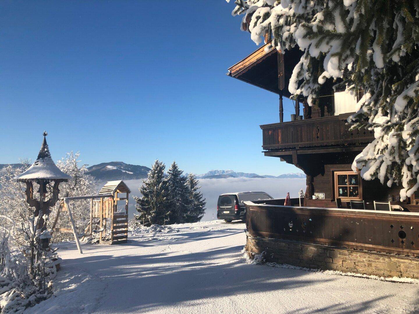 Winterlandschaft mit verschneitem Chalet, Spielplatz und Bergblick über eine Nebeldecke.