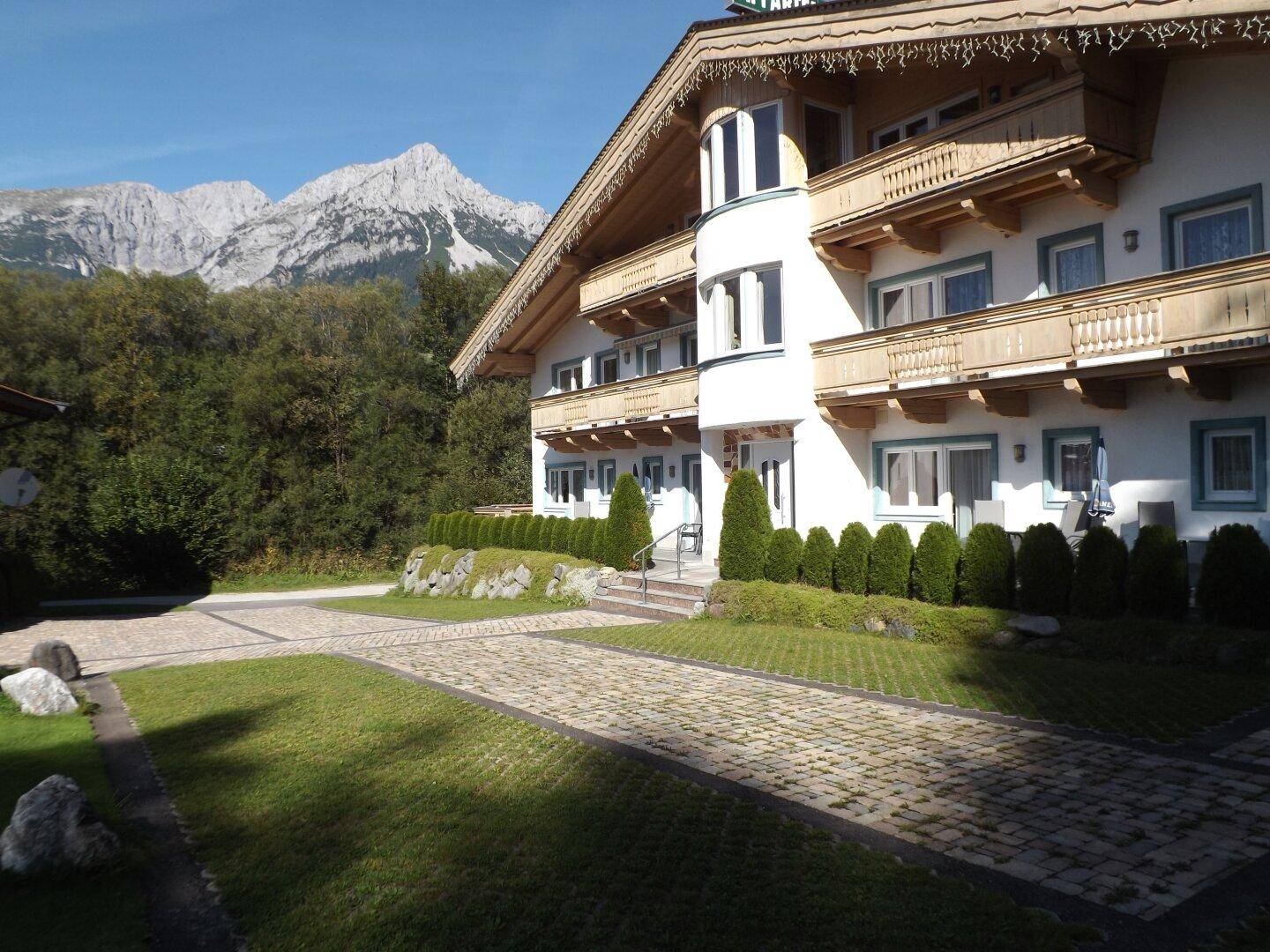 Alpines Gebäude mit Holzbalkonen und gepflasterter Auffahrt vor einer Bergkulisse.