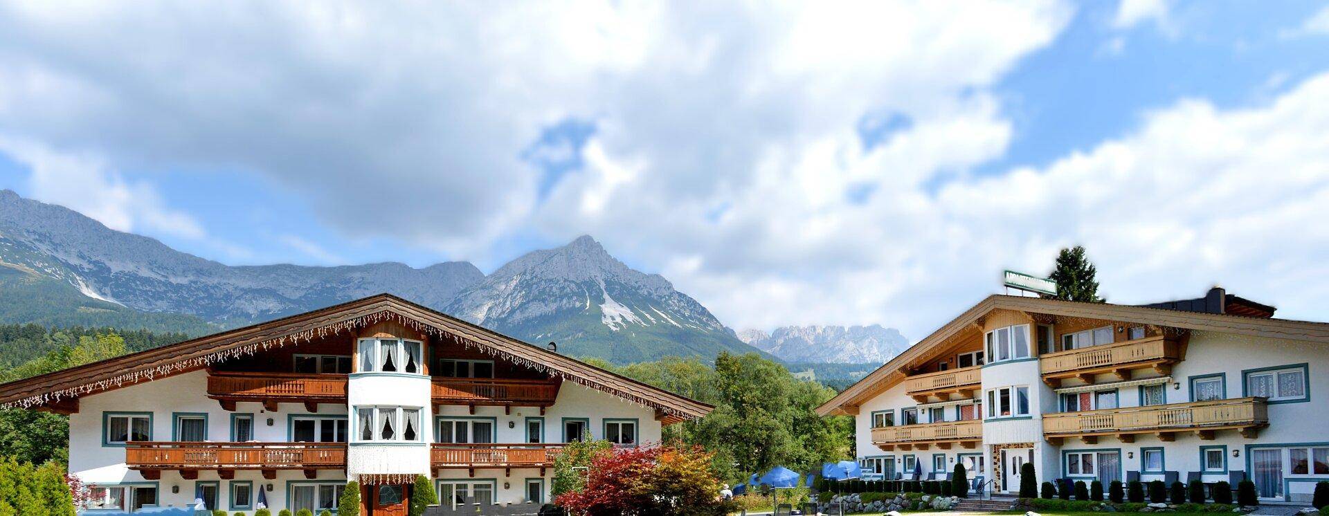 Zwei traditionelle Gebäude im alpinen Stil mit Holzbalkonen vor einer beeindruckenden Berglandschaft.