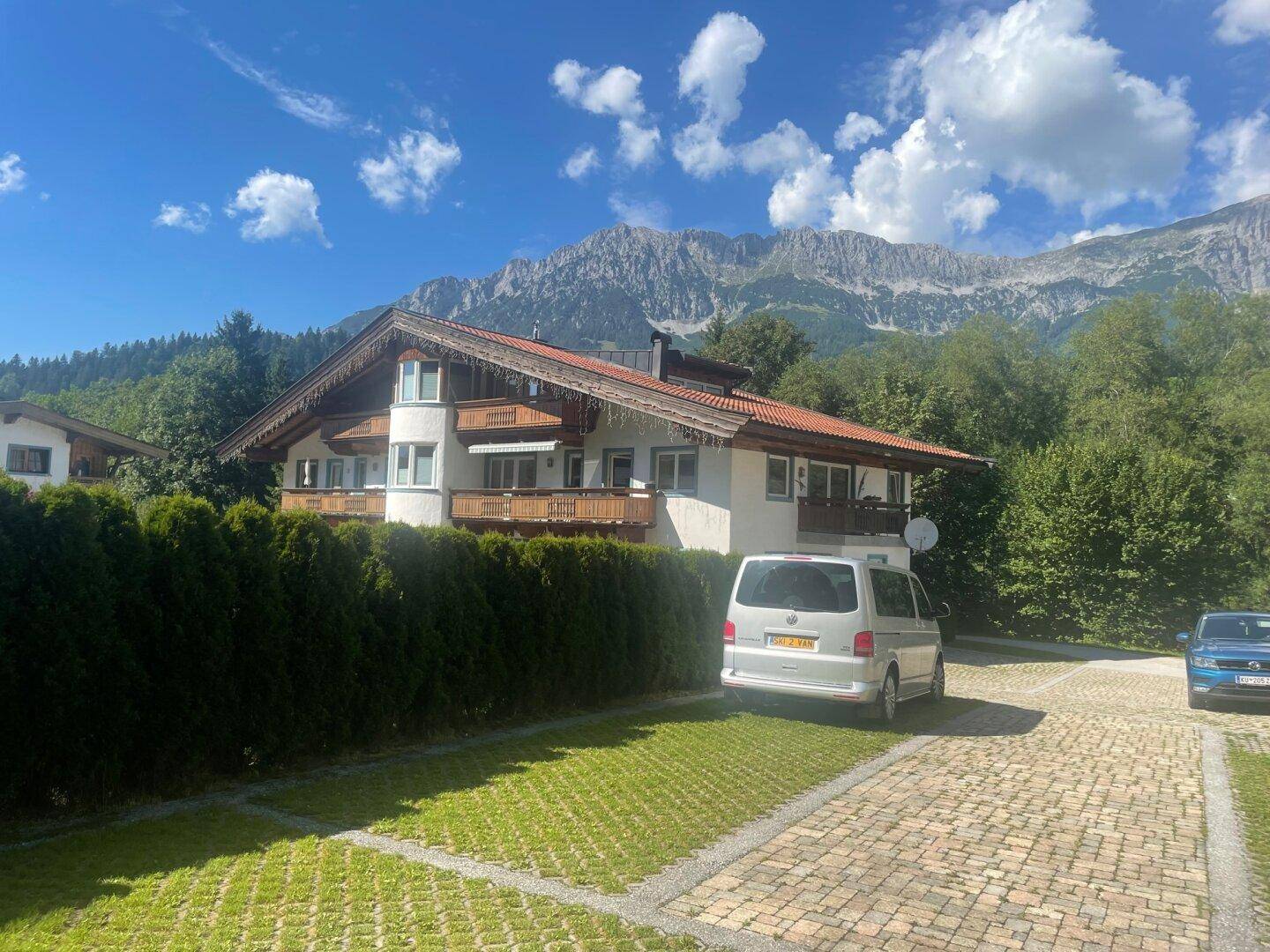 Alpines Gebäude mit Holzbalkonen und Parkplätzen vor einer malerischen Bergkulisse.