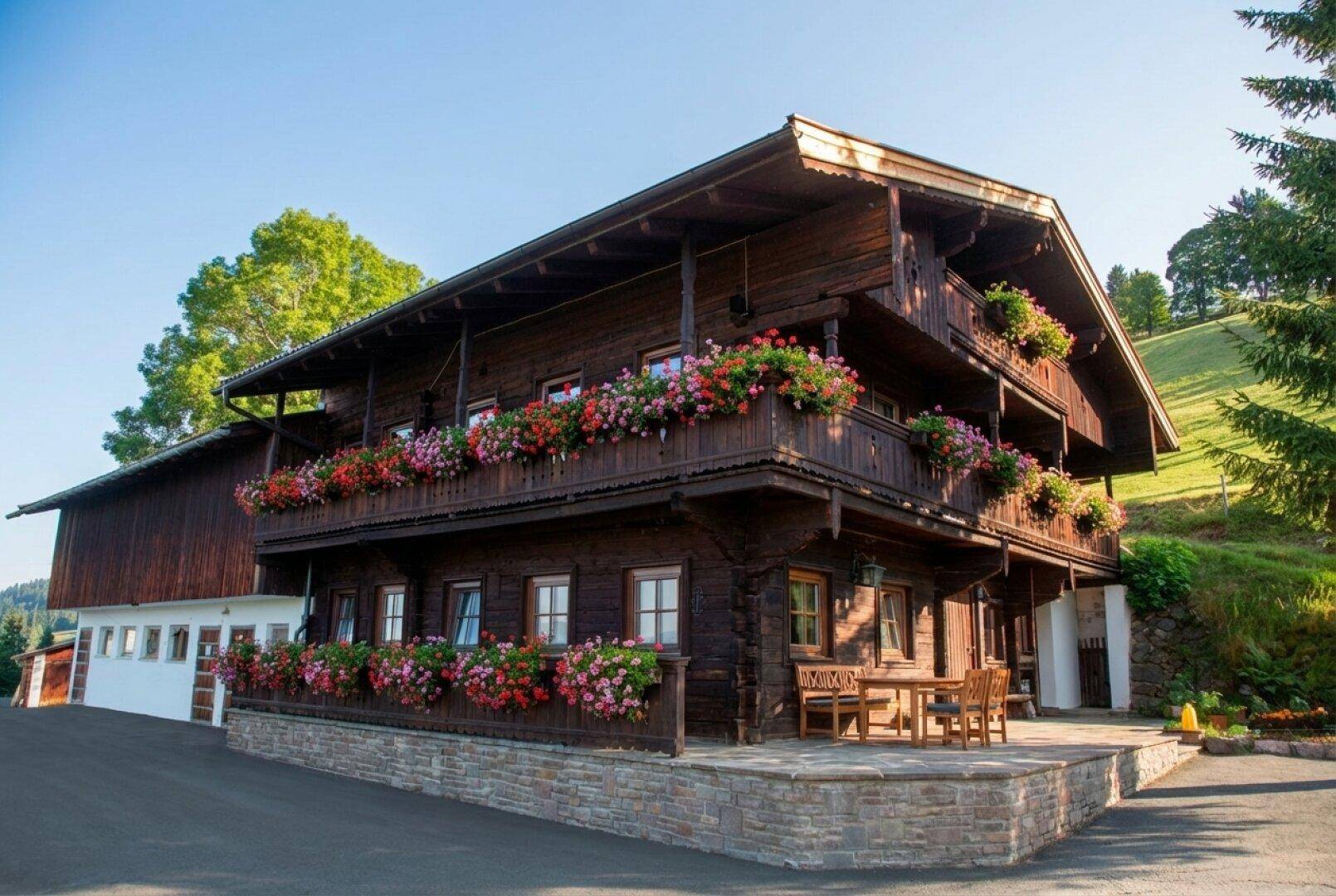 Traditionelles Holzhaus mit blühenden Blumenkästen und gemütlicher Terrasse im Sommer, umgeben von grünen Hügeln.