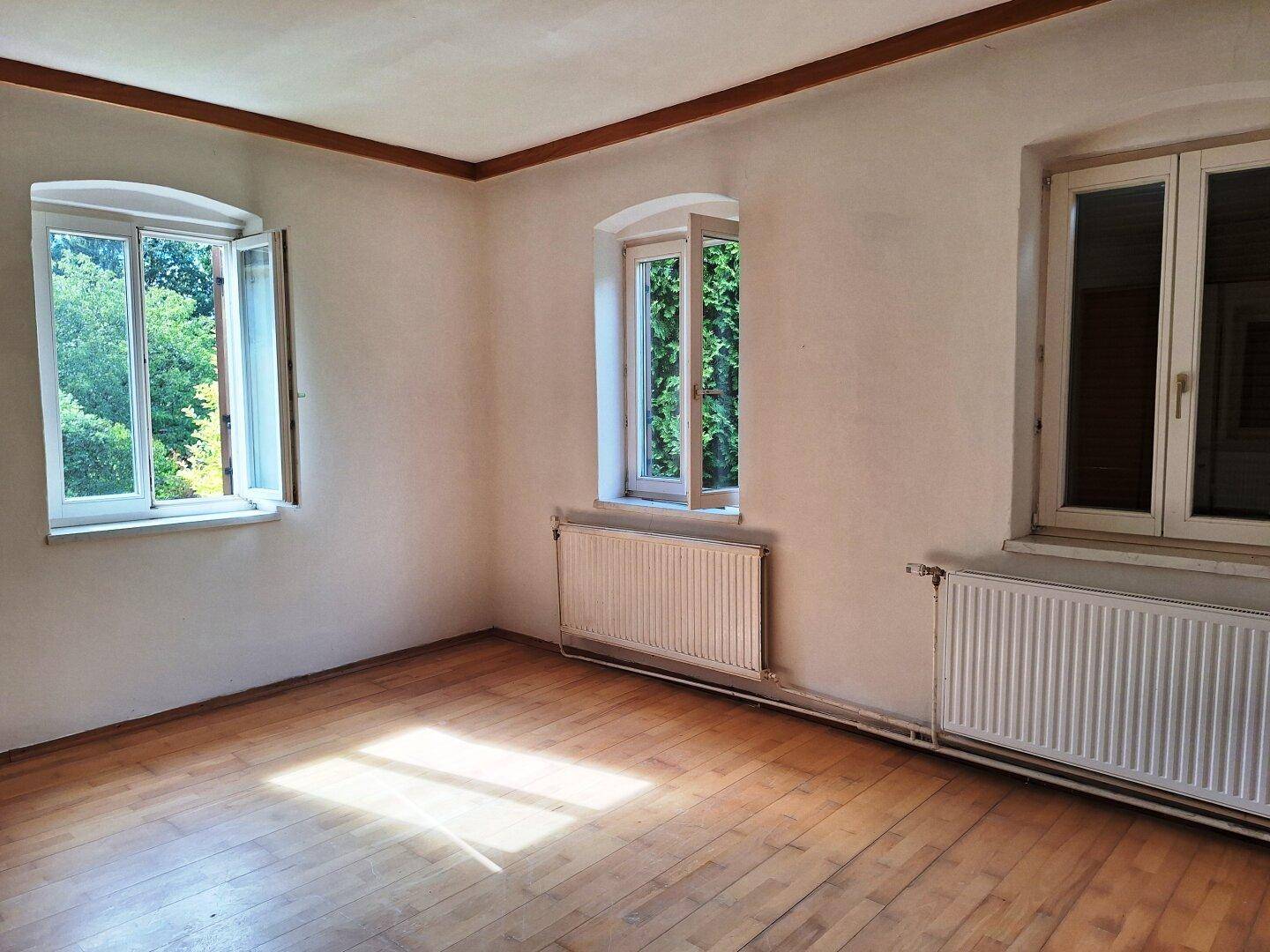 Leerer Raum mit Holzboden, weißen Wänden und mehreren Fenstern, die viel Licht hereinlassen.