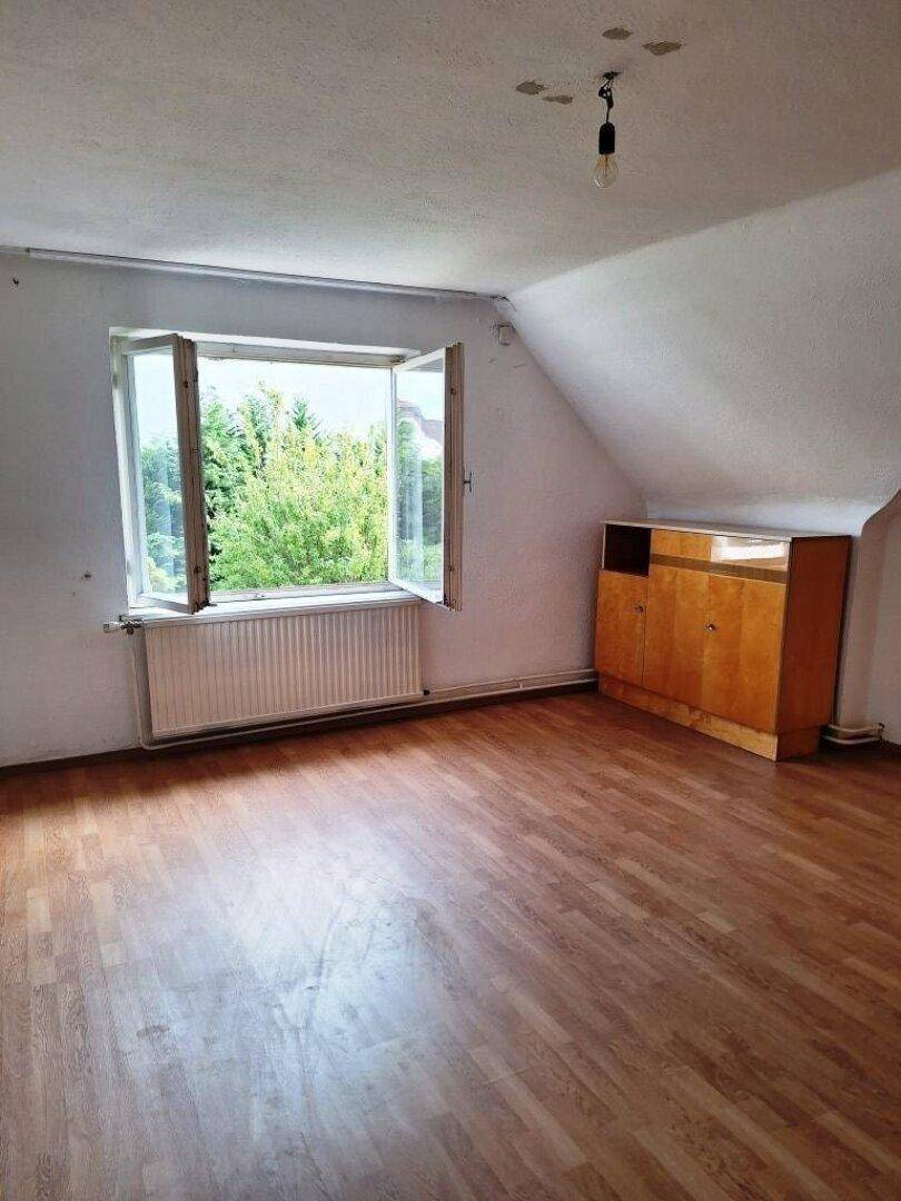Leeres Dachgeschosszimmer mit Laminatboden, Fenster und einem kleinen Holzschrank.