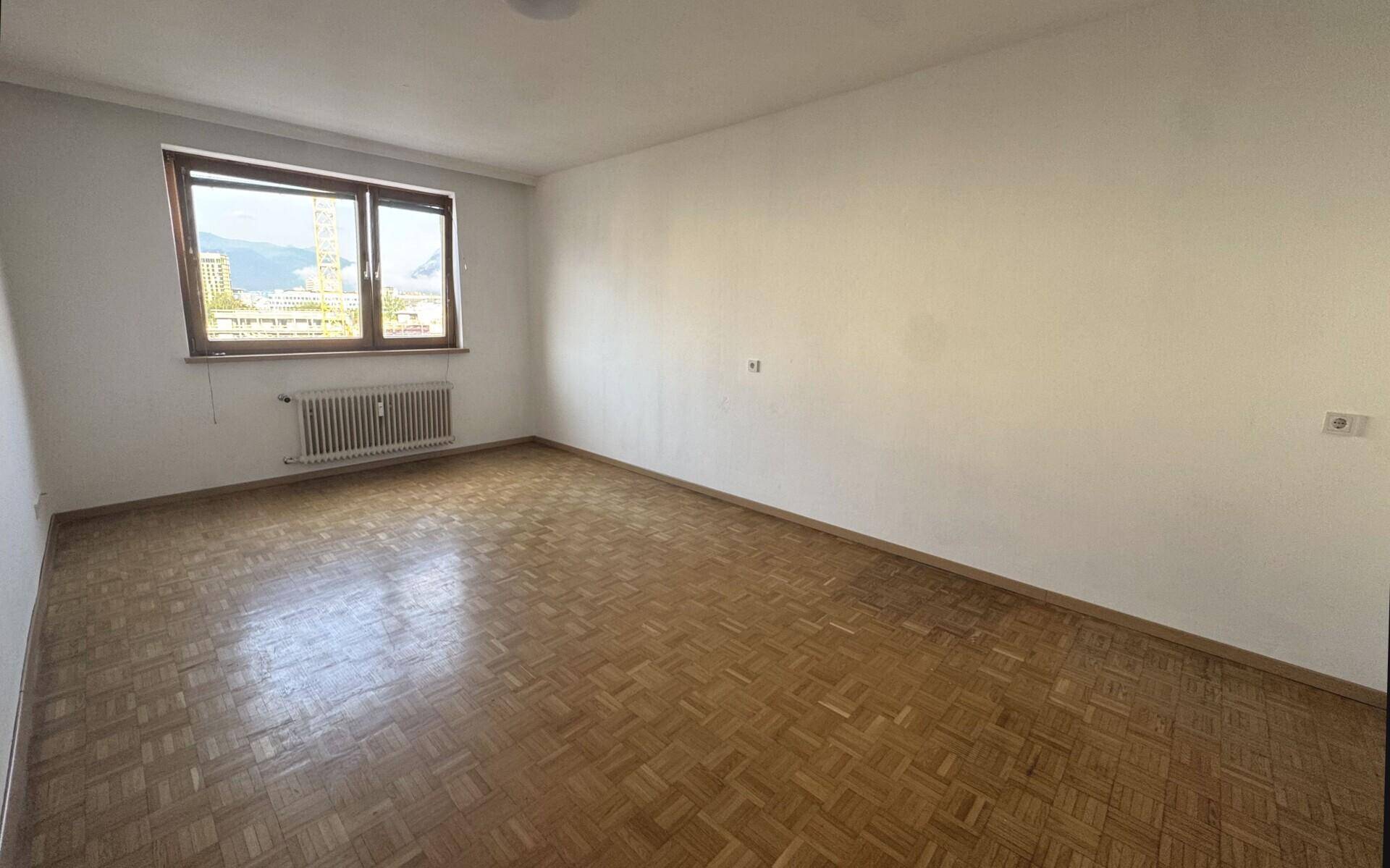 Heller Raum mit klassischem Parkettboden und einem Fenster, das den Blick nach draußen freigibt.